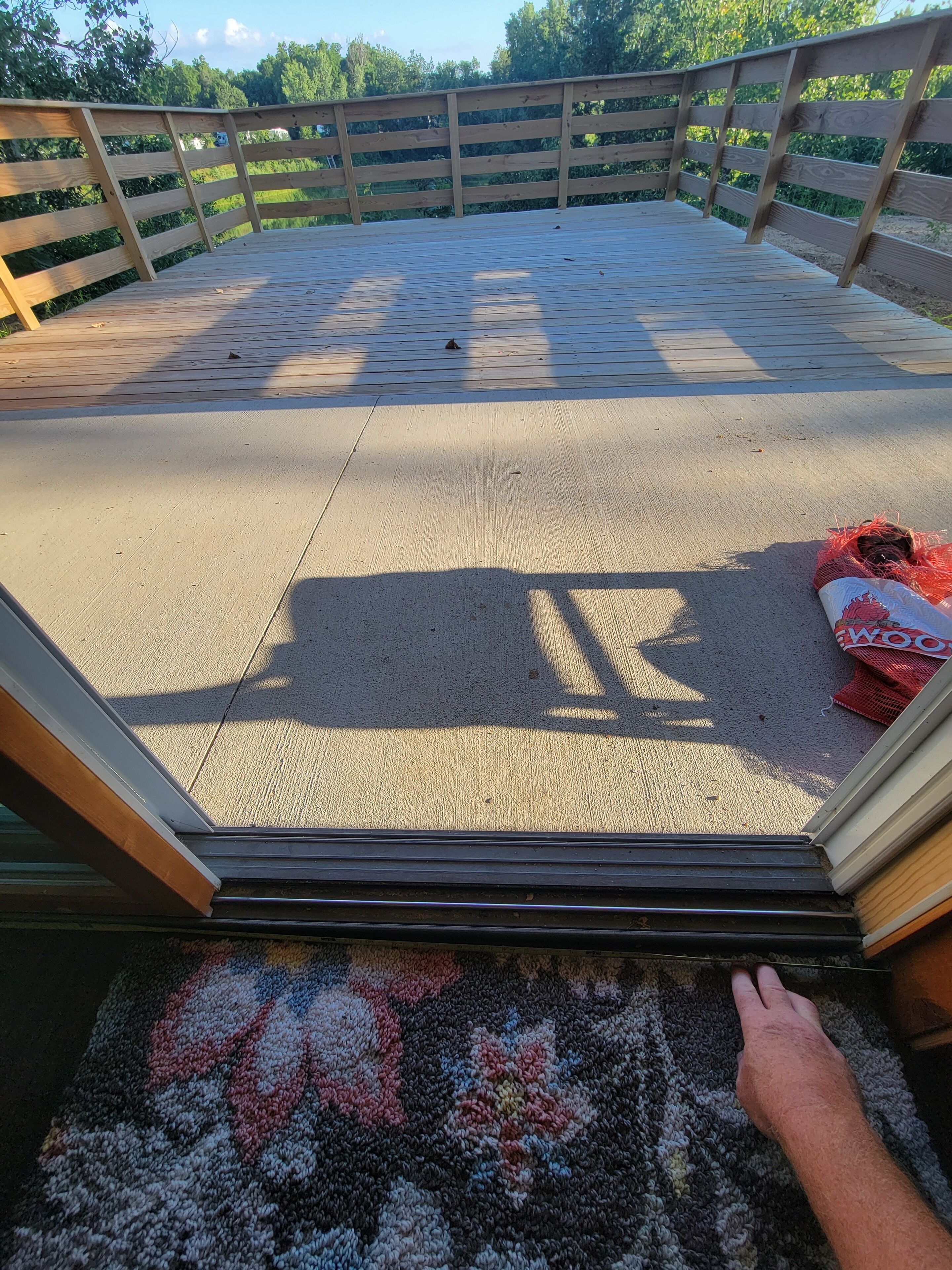 Une porte coulissante ouverte donne sur une terrasse dotée d’une rambarde en bois, d’un sol en béton et d’un sac rouge posé par terre.
