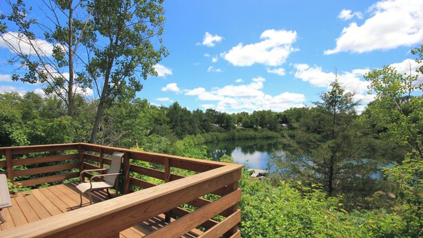 A wooden deck with a chair and a table, overlooking a lake surrounded by greenery. - Quaint Lakeview Cabin  (Wadena)