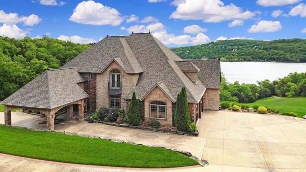 A large, multi-story stone house with a prominent gabled roof, a covered patio, and a view of a lake and forested hills. - Luxurious Spacious Chateau on Table Rock Lake with Hot Tub (Ridgedale)