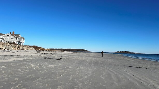 Une plage avec une grande maison sur la gauche, une personne qui marche sur le sable et un ciel bleu dégagé.