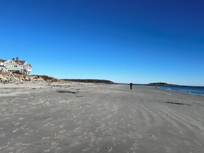 Een strand met een groot huis aan de linkerkant, een persoon die over het zand loopt, en een helderblauwe lucht.