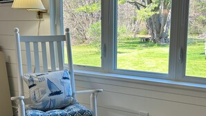 Une chaise blanche avec des coussins à motifs, placée près d’une fenêtre donnant sur une cour verdoyante et des arbres.