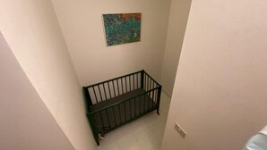 Un coin de pièce avec un berceau en métal vert adossé à un mur blanc, un petit tableau au-dessus et un luminaire fixé au mur.