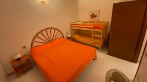 Une petite pièce, de taille modeste, comprenant un lit simple, un lit superposé, une armoire en bois, une petite table avec une lampe et un luminaire fixé au mur.