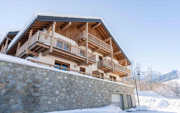 A multi-story chalet with wooden balconies and stone walls, set against a snowy mountain backdrop. - 380m from shops, 1st floor, view forest, terrace, parking, ski locker, 40m², Valmorel (Les Avanchers-Valmorel)