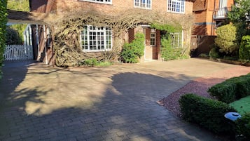 A brick house with a paved driveway, a white fence, and a garden area with bushes and a small tree.
