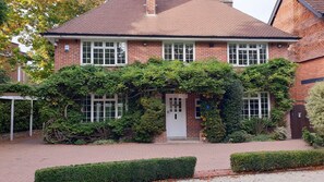 A two-story brick house with a white door and windows, surrounded by lush greenery and a well-maintained garden.