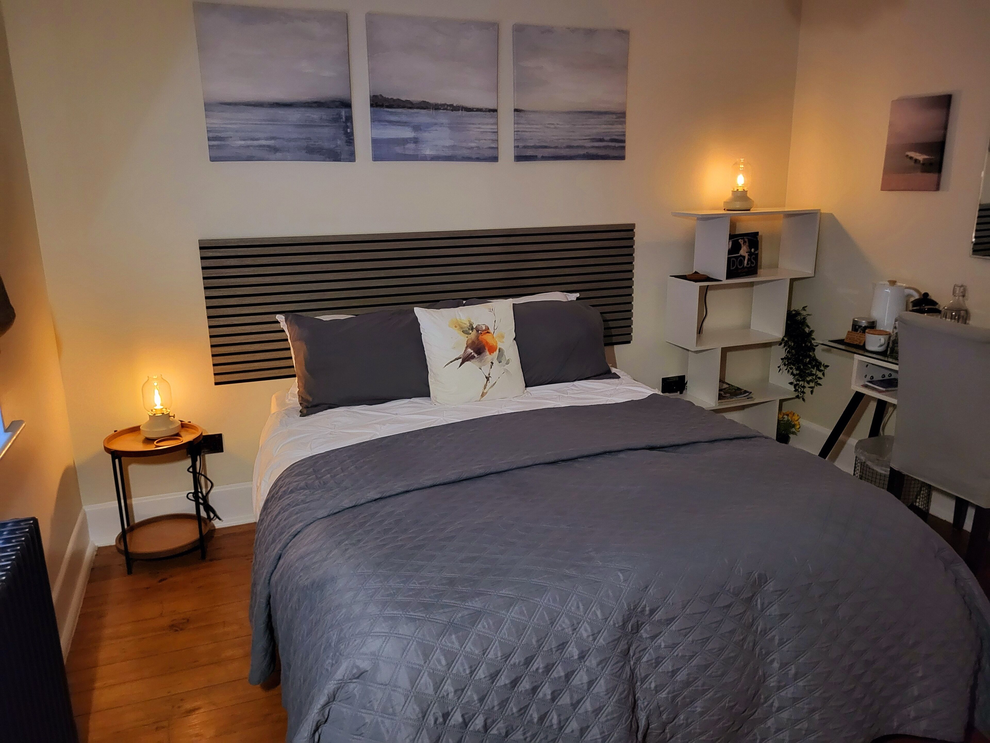 A bedroom with a large bed, a bedside table with a lamp, a nightstand, a dresser, and a wall-mounted shelf with a lamp.