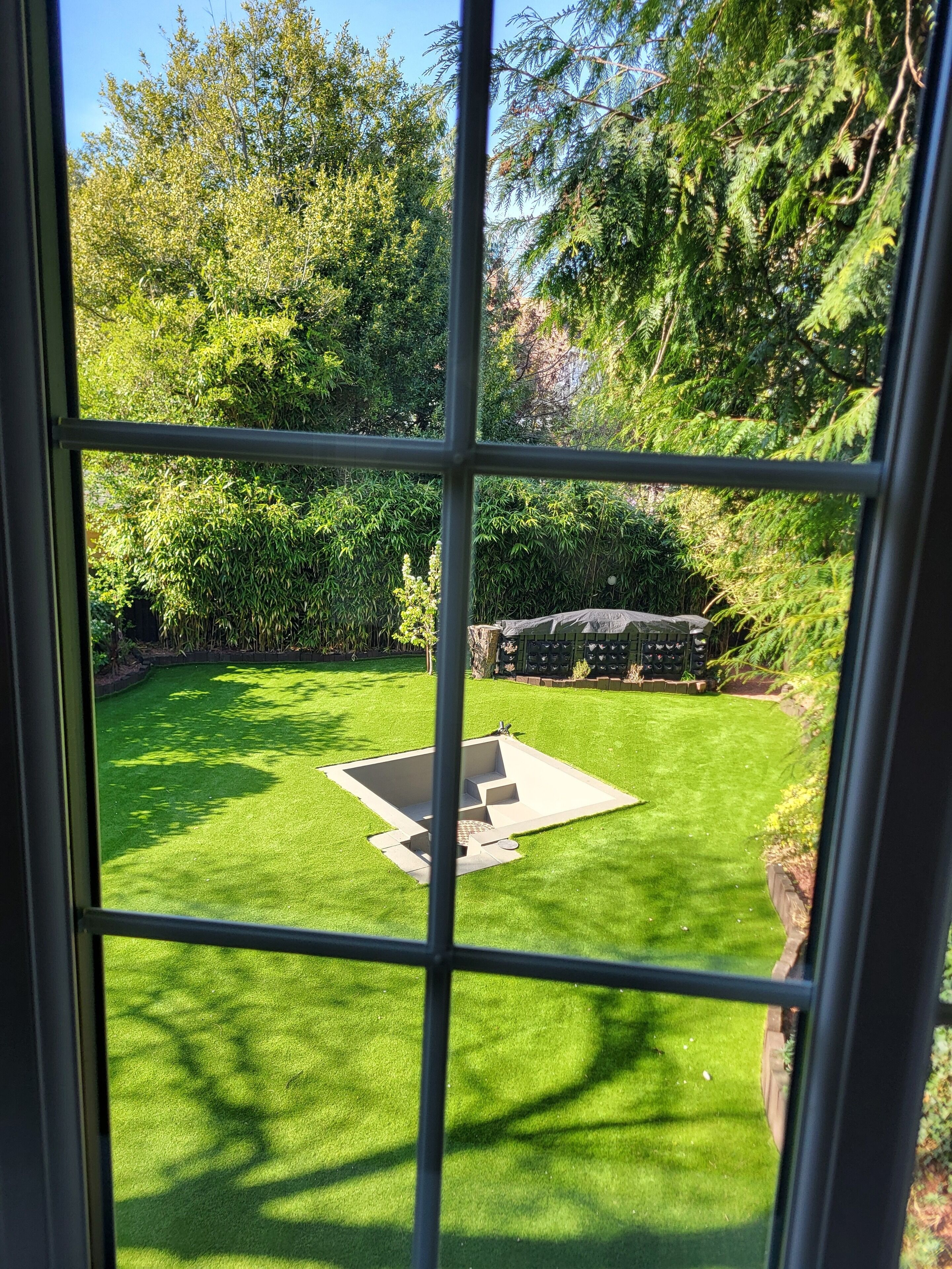 A view through a window of a well-maintained garden with a central lawn area, surrounded by dense foliage and trees.