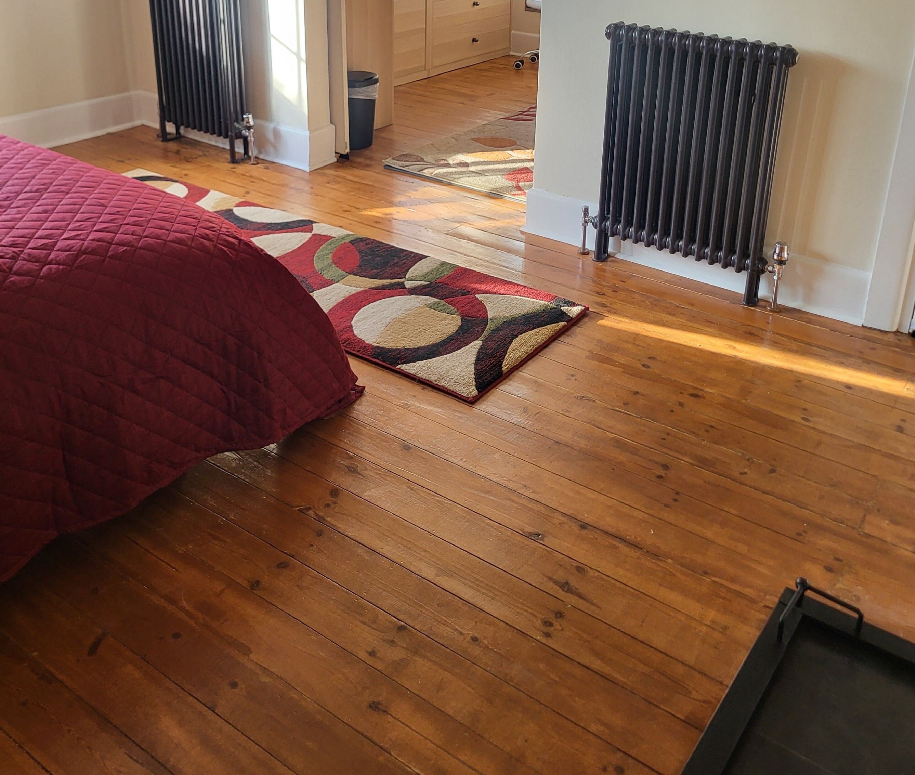 Un dormitorio con piso de madera, cubrecama rojo y alfombra estampada.