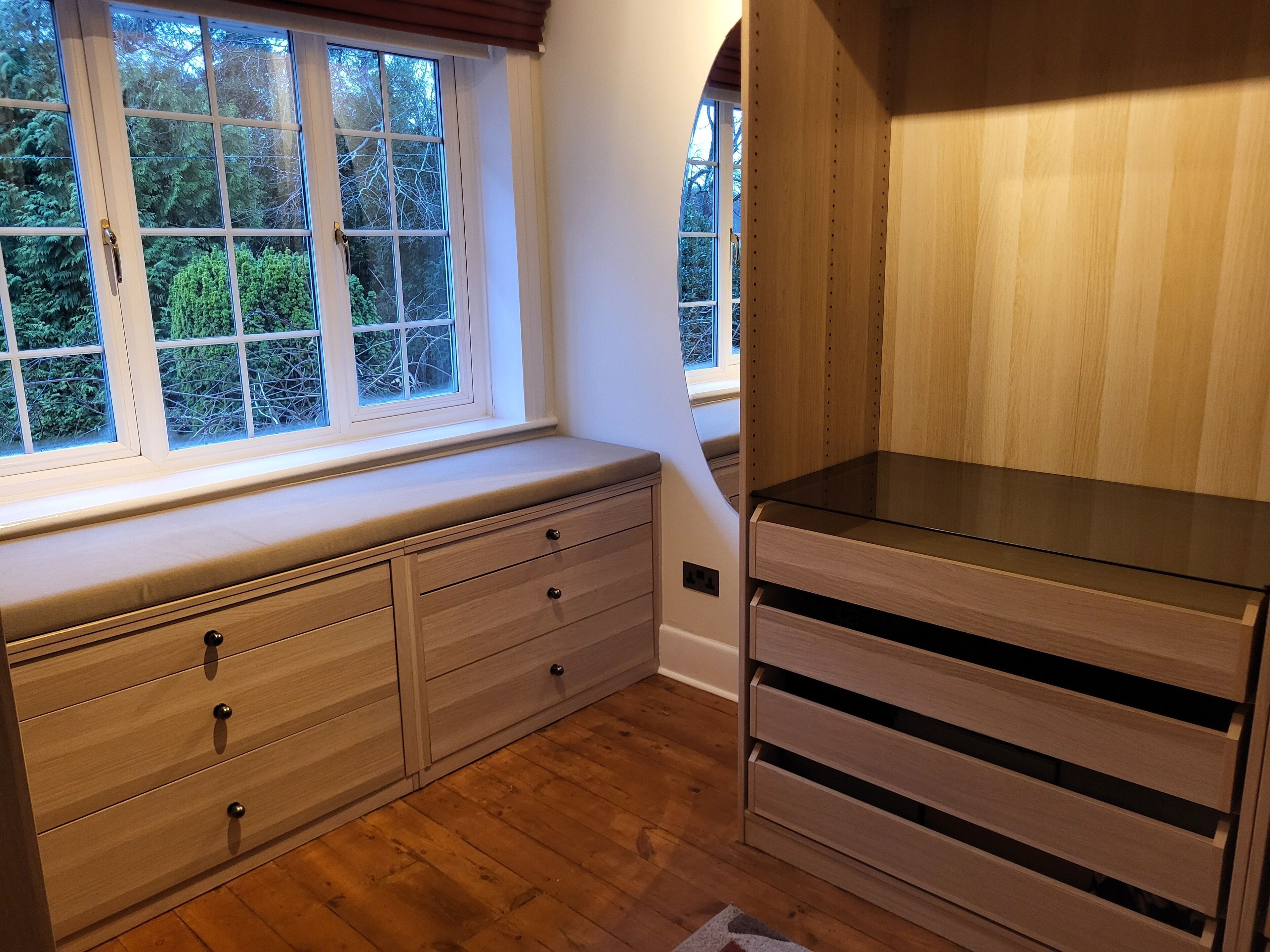 A room with a built-in bench along a window, wooden flooring, and a built-in cabinet with drawers.