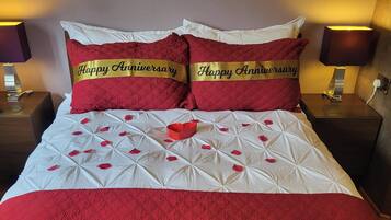 Una cama con colcha blanca y roja, dos almohadas con el texto 'Happy Anniversary' y pétalos de rosa.