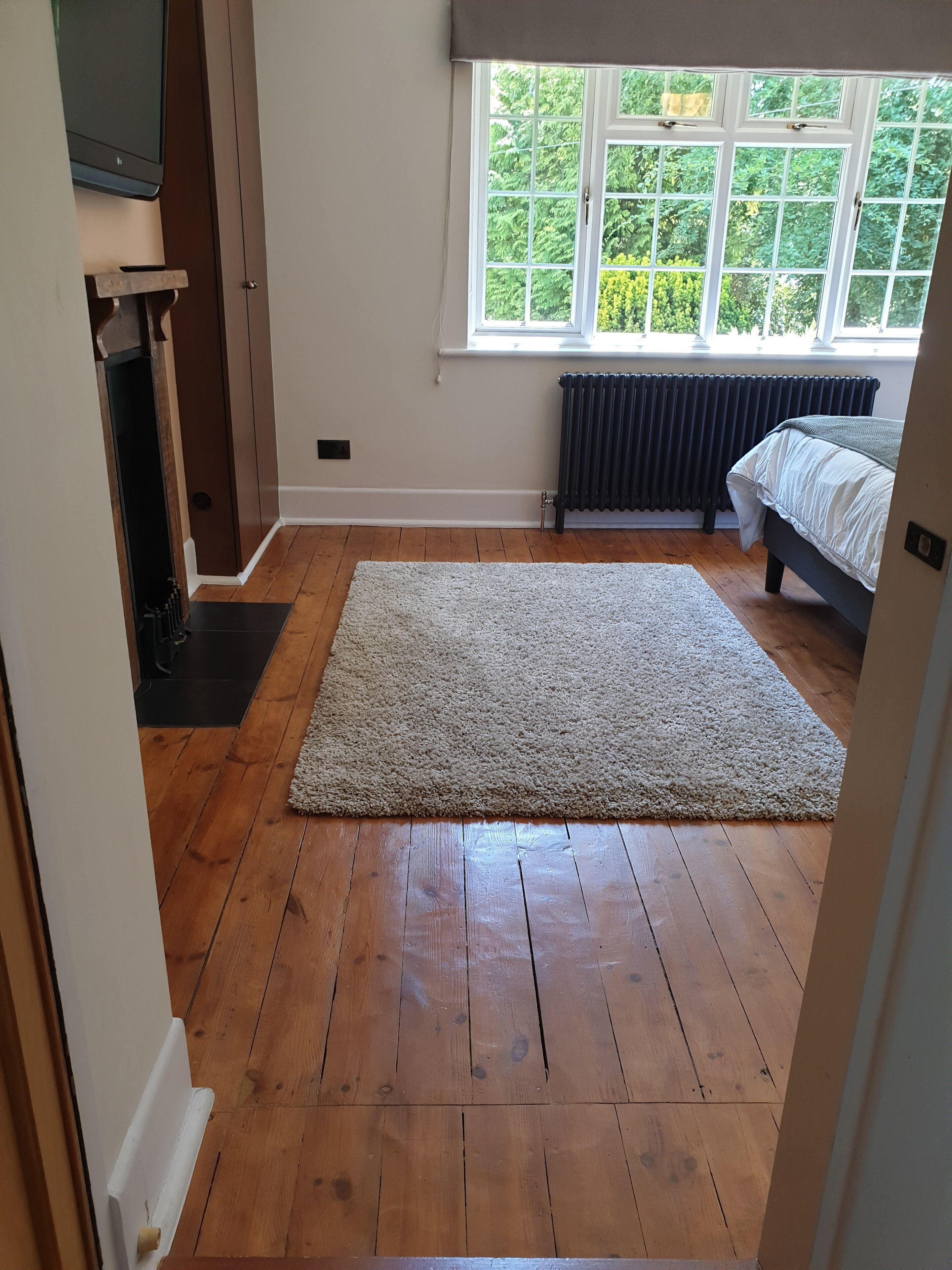 A bedroom with a wooden floor, a large rug, a bed, a radiator, and a window with a view of greenery.