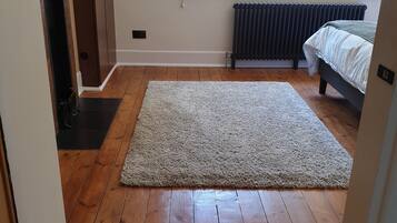 A bedroom with a wooden floor, a large rug, a bed, a radiator, and a window with a view of greenery.
