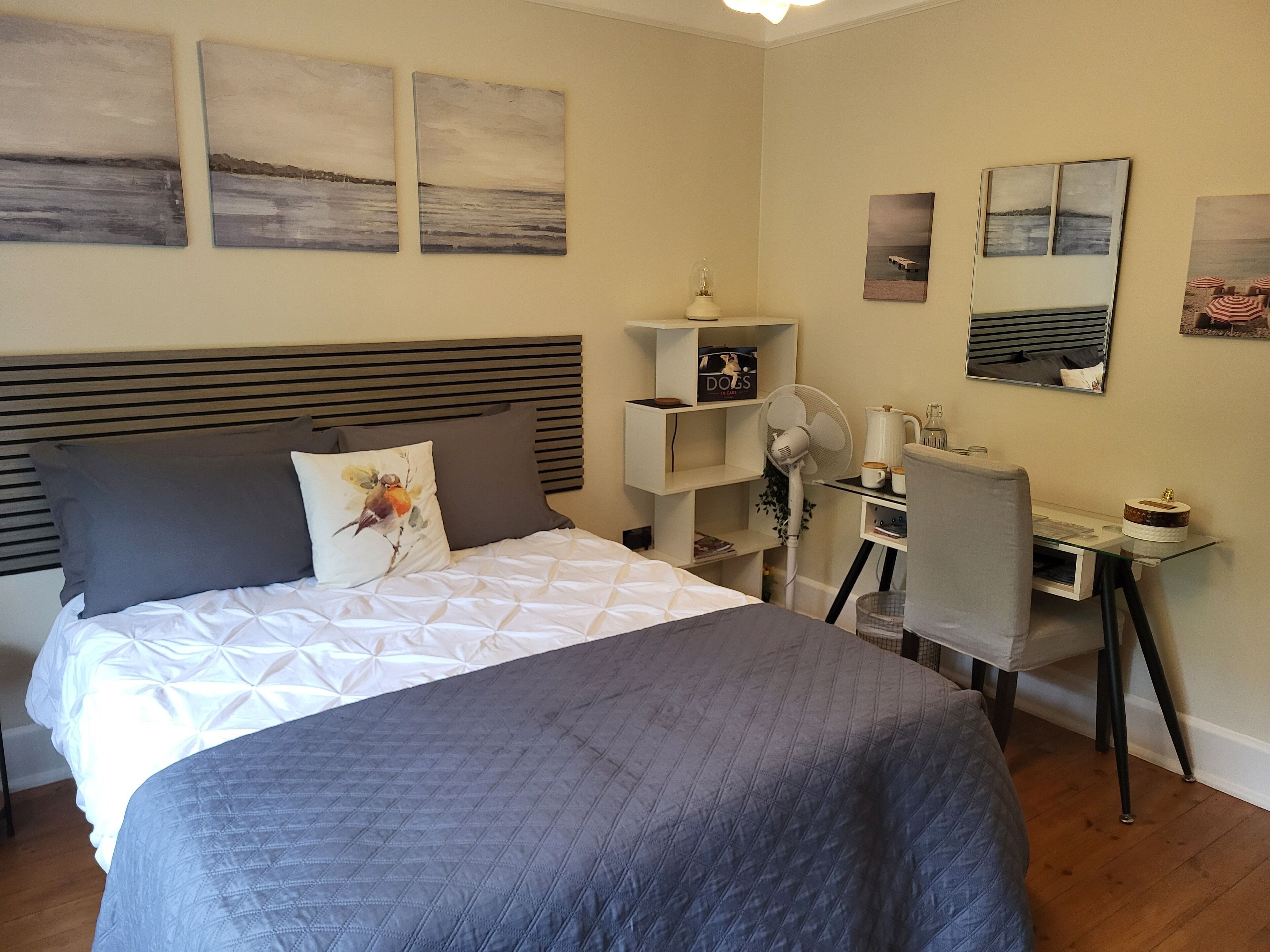 Un dormitorio ordenado con una cama grande, un escritorio y una silla. Hay obras de arte en las paredes y un pequeño estante con objetos.