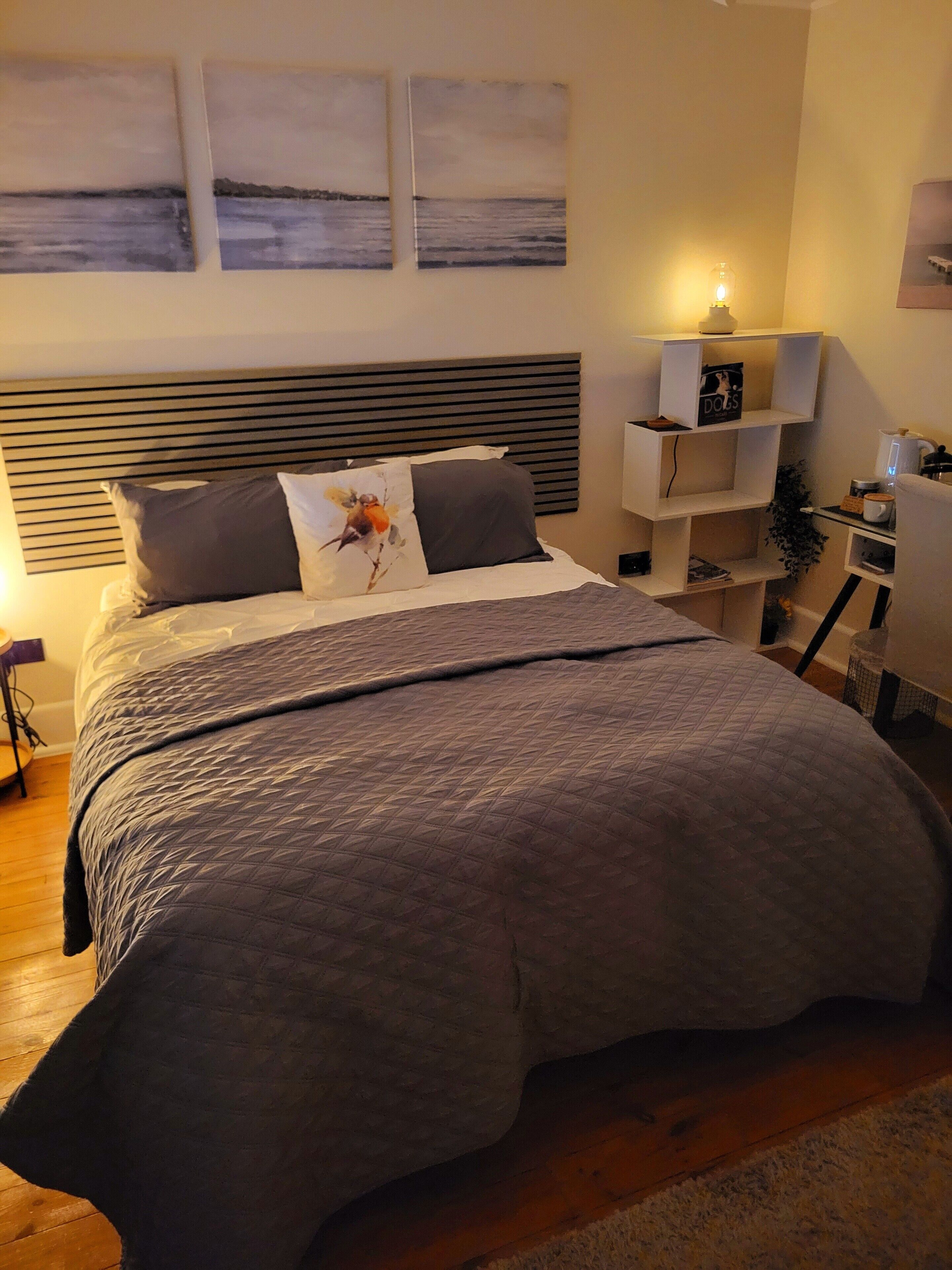 A modern bedroom with a large bed, a bedside table with a lamp, and three wall-mounted paintings.