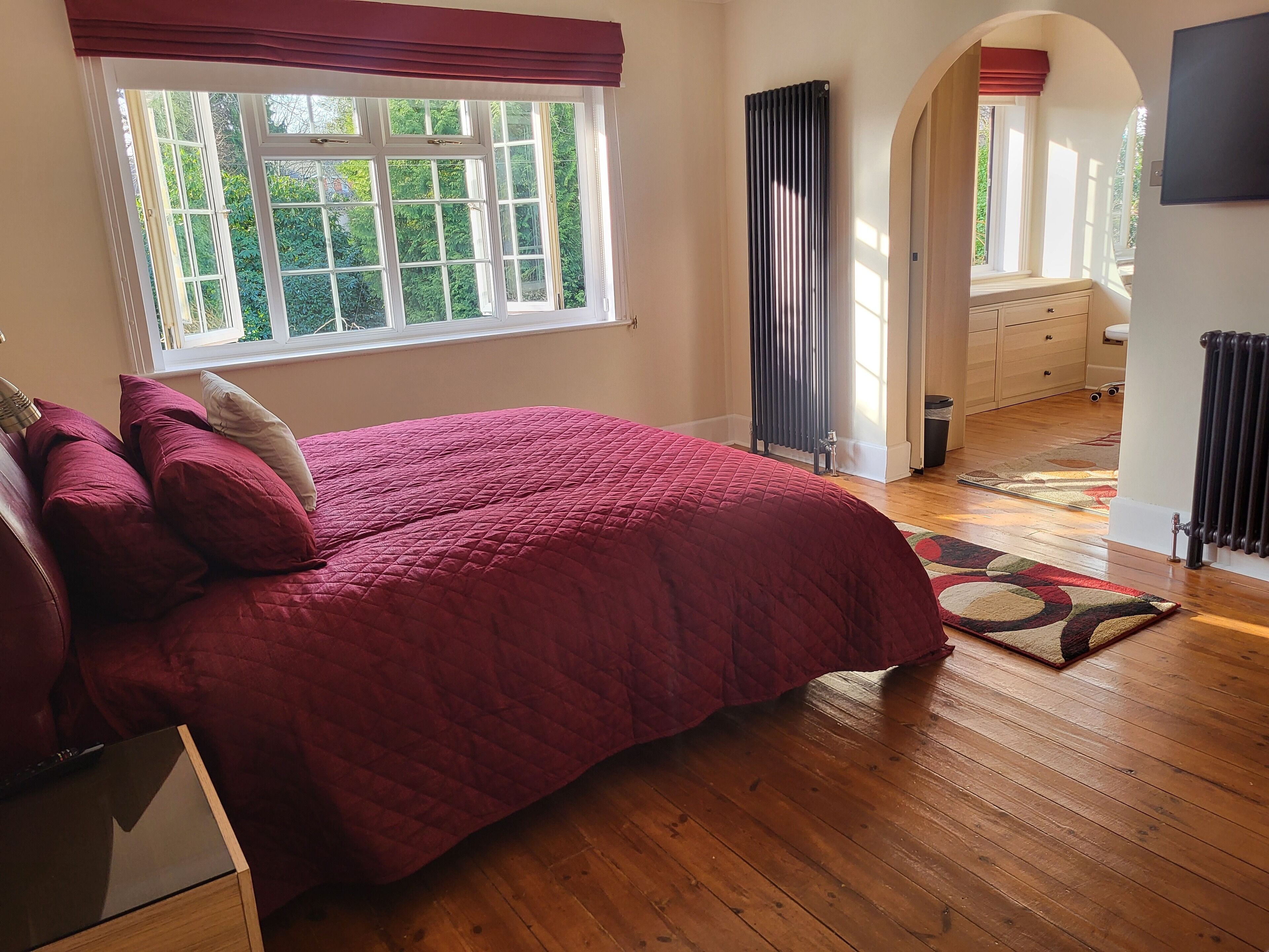 Un dormitorio con una cama grande, suelo de madera, una ventana con cortinas, un radiador y vistas a un jardín.