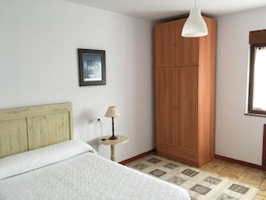 A bedroom with a bed, a wooden wardrobe, a small table with a lamp, a framed picture on the wall, and a window with a view.