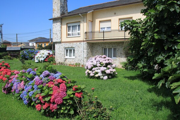 A two-story house with a stone facade, a balcony with a black railing, and a well-maintained garden with various flowers.