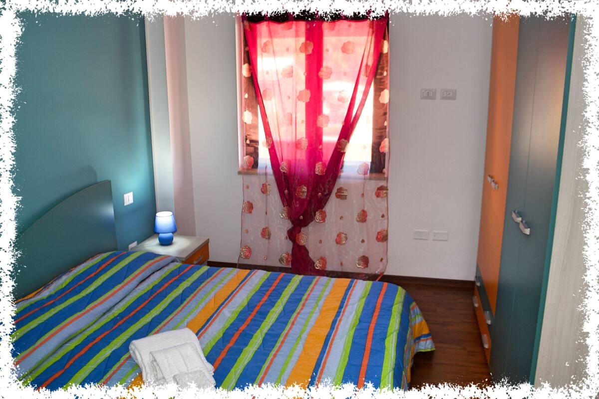 A bedroom with a bed, a nightstand, a lamp, a window with sheer curtains, and a door.