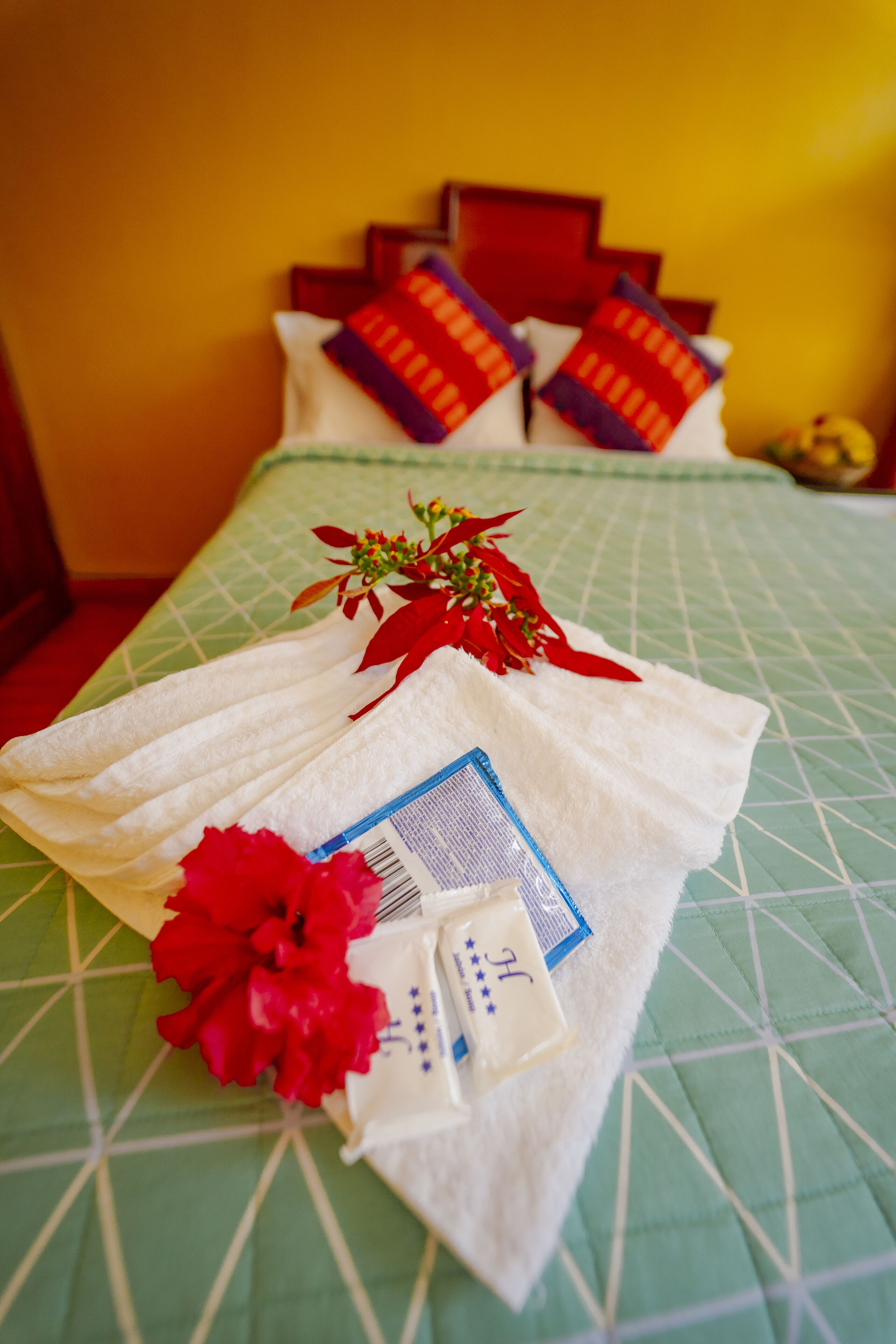 A neatly made bed with a green quilt, red and white pillows, a red flower, and a folded towel with a card.