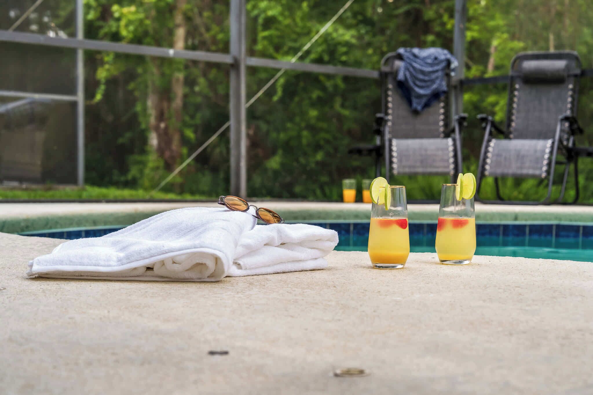 A poolside area with a towel, sunglasses, and two glasses of a yellow drink with a red garnish.