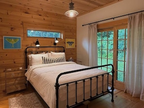 A wooden cabin bedroom with a metal bed frame, a nightstand, a window with curtains, and a ceiling light.