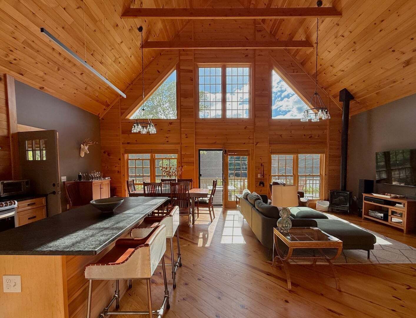 A spacious log cabin interior with a kitchen, dining area, and living room, featuring large windows and a high ceiling.