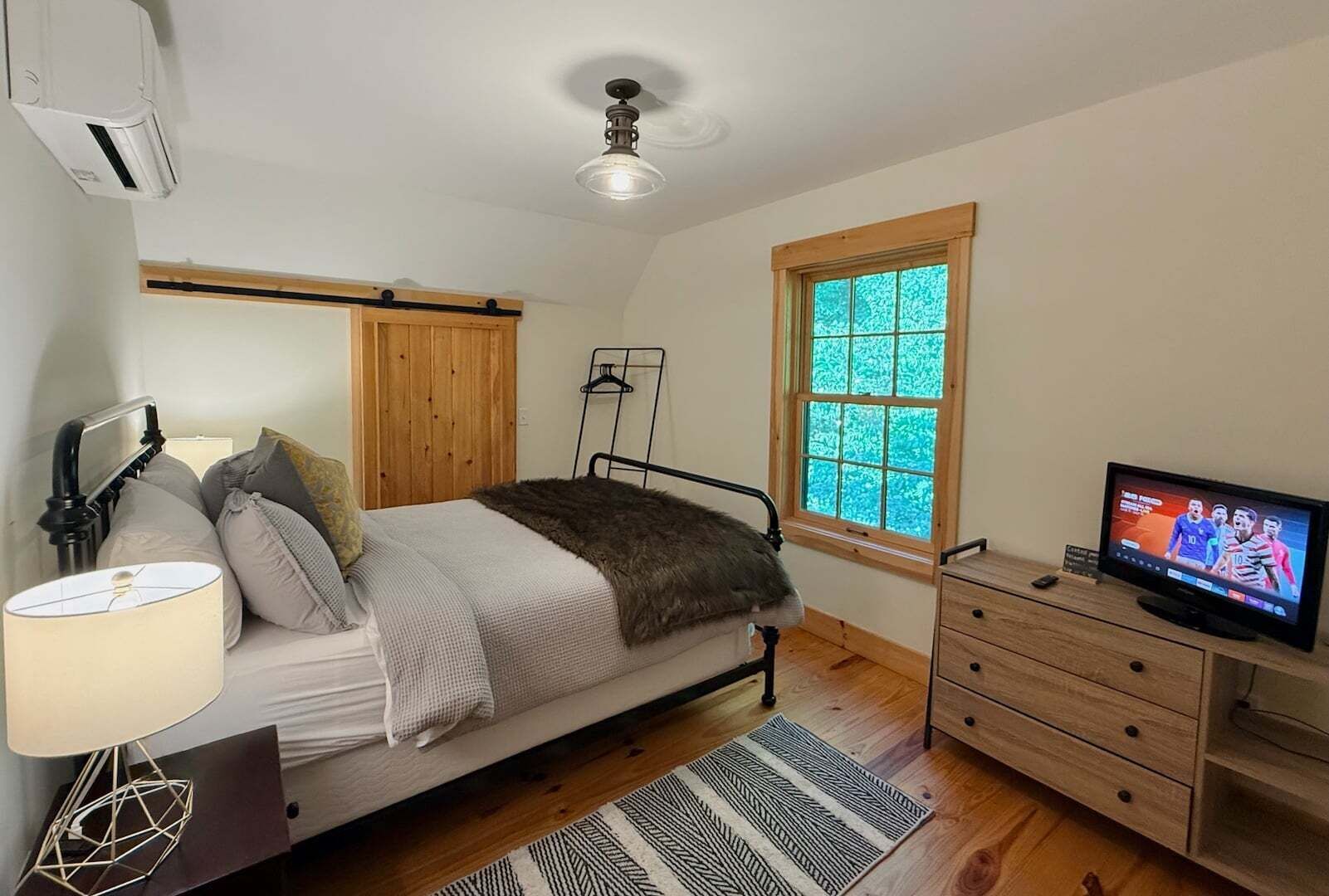 A bedroom with a bed, bedside lamp, wooden wardrobe, TV, and a window with a view.