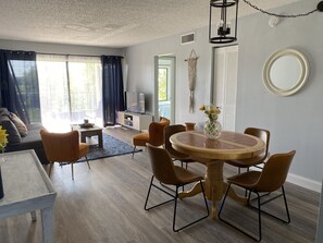 A modern living room with a round wooden dining table surrounded by brown chairs, a sofa, and a TV on a white cabinet.