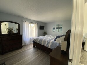 A bedroom with a wooden dresser, a bed with patterned bedding, a large mirror, and a TV on the wall.