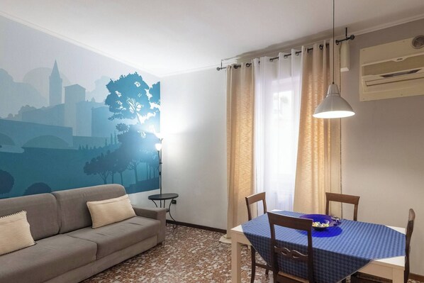 A living room with a sofa, a dining table with chairs, a wall mural, a lamp, and an air conditioning unit.