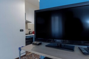 A modern room with a large flat-screen TV on a stand, a small desk with a keyboard, and a kitchenette in the background.