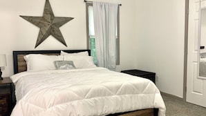 Un lit bien fait, avec une literie blanche, une décoration en forme d’étoile accrochée au mur, une table de chevet avec une lampe et une fenêtre avec des rideaux.