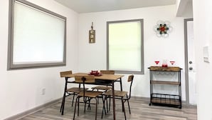 Un coin repas avec une table et des chaises en bois, deux grandes fenêtres équipées de stores, un panneau lumineux indiquant «repas» fixé au mur, et une petite étagère avec des objets rouges.