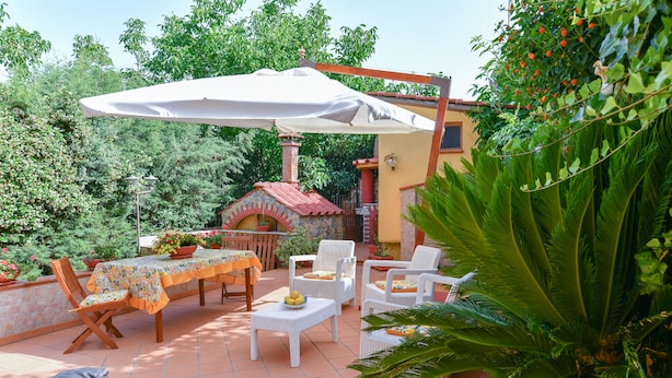 Une terrasse extérieure avec une table, des chaises et un grand parasol. On aperçoit au fond un petit four en briques et des plantes en pot.