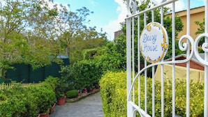 Un portail en fer forgé, sur lequel on peut lire l’inscription « Daisy House », mène à un sentier bordé d’une végétation luxuriante et de plantes en pot.