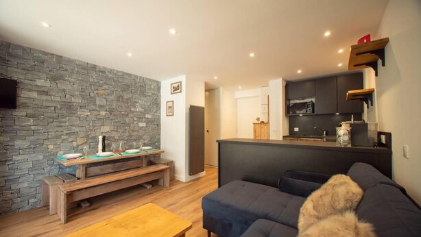 A modern living room with a stone wall, a dark sofa, a wooden coffee table, and a kitchen area with a microwave and cabinets. - 50m from ski slopes, 1st floor, view mountain, balcony, tv, ski locker, 34m², Valmorel (Valmorel)