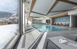 A modern indoor swimming pool with clear glass walls, lounge chairs, and a view of a snowy landscape.