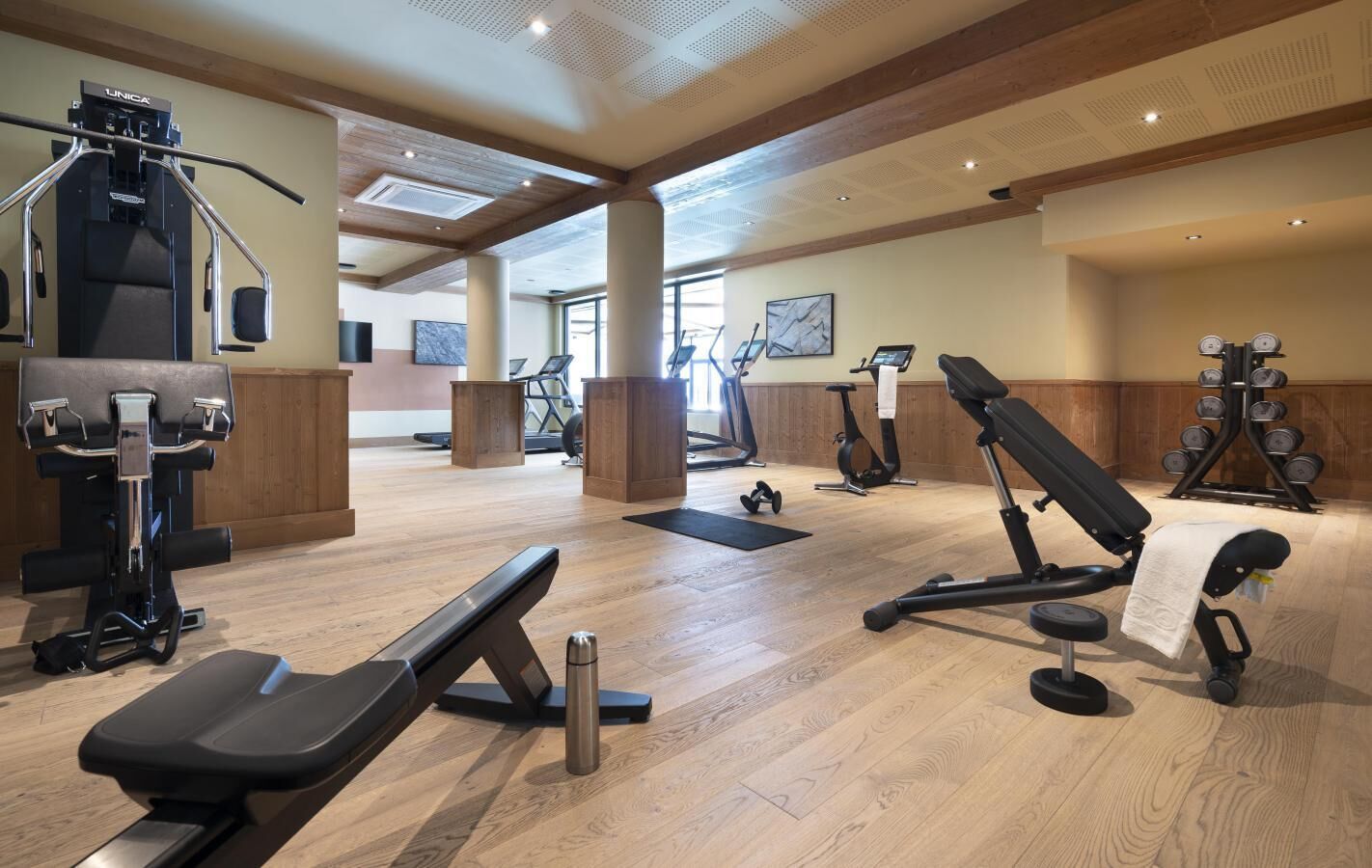 A gym with various exercise equipment, wooden flooring, and large windows allowing natural light.
