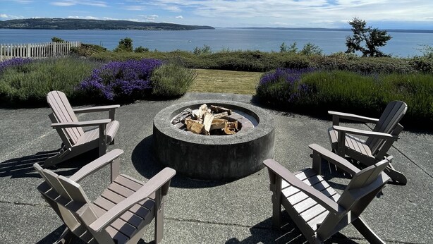 Un espace extérieur aménagé avec des chaises Adirondack disposées autour d’un foyer, offrant une vue panoramique sur un lac et des collines lointaines.