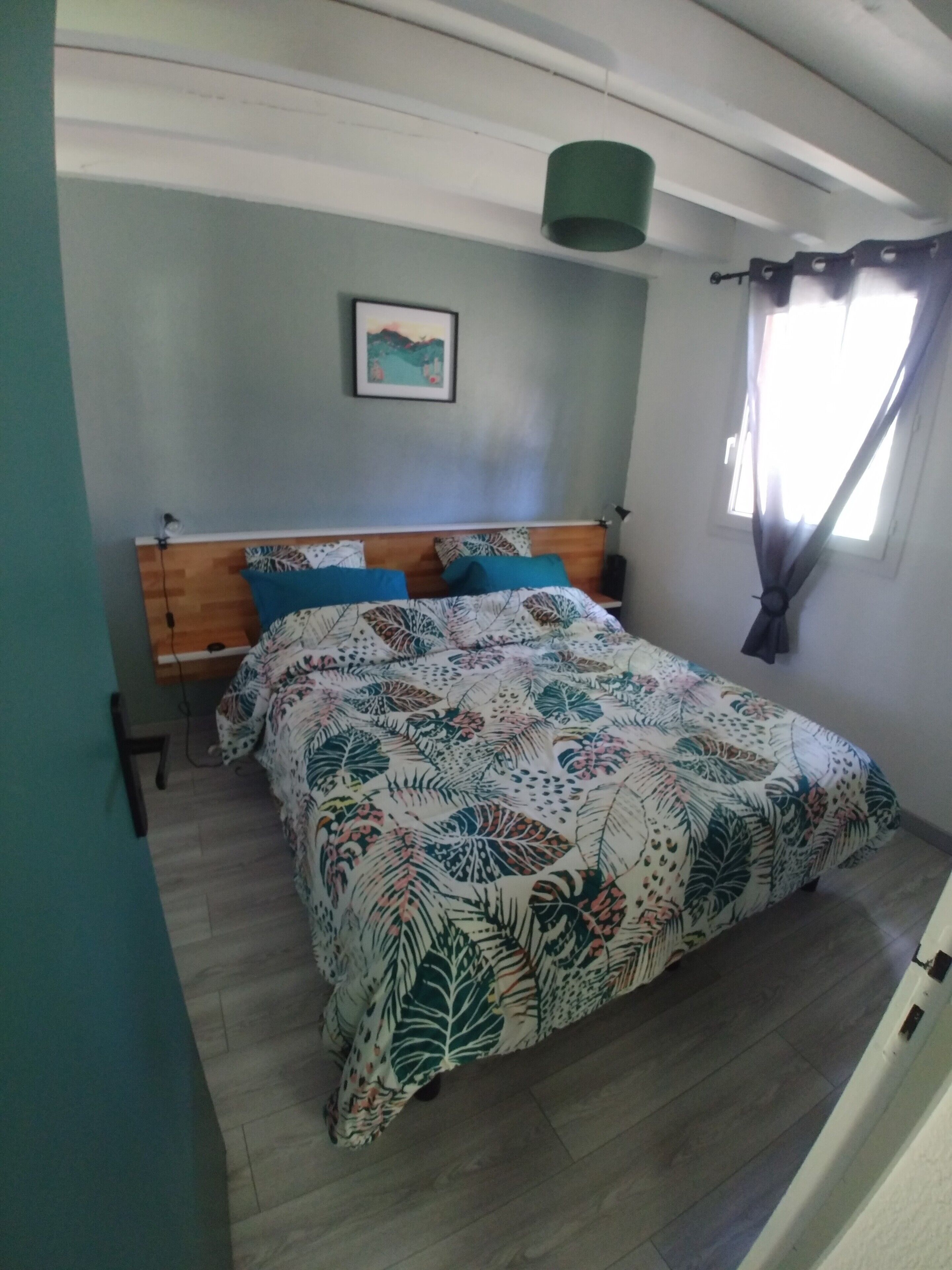 Une chambre à coucher avec une tête de lit en bois, un lit recouvert d’une couvre-lit à motifs floraux, une table de chevet, une lampe fixée au mur, un tableau et une fenêtre avec des rideaux.