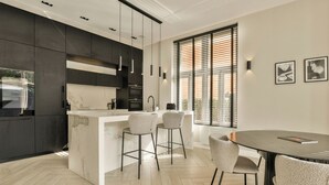 A modern kitchen with dark cabinetry, a marble island, and bar stools. There is a dining area with a round table and chairs. - Luxury Rijksmuseum house (Amsterdam)