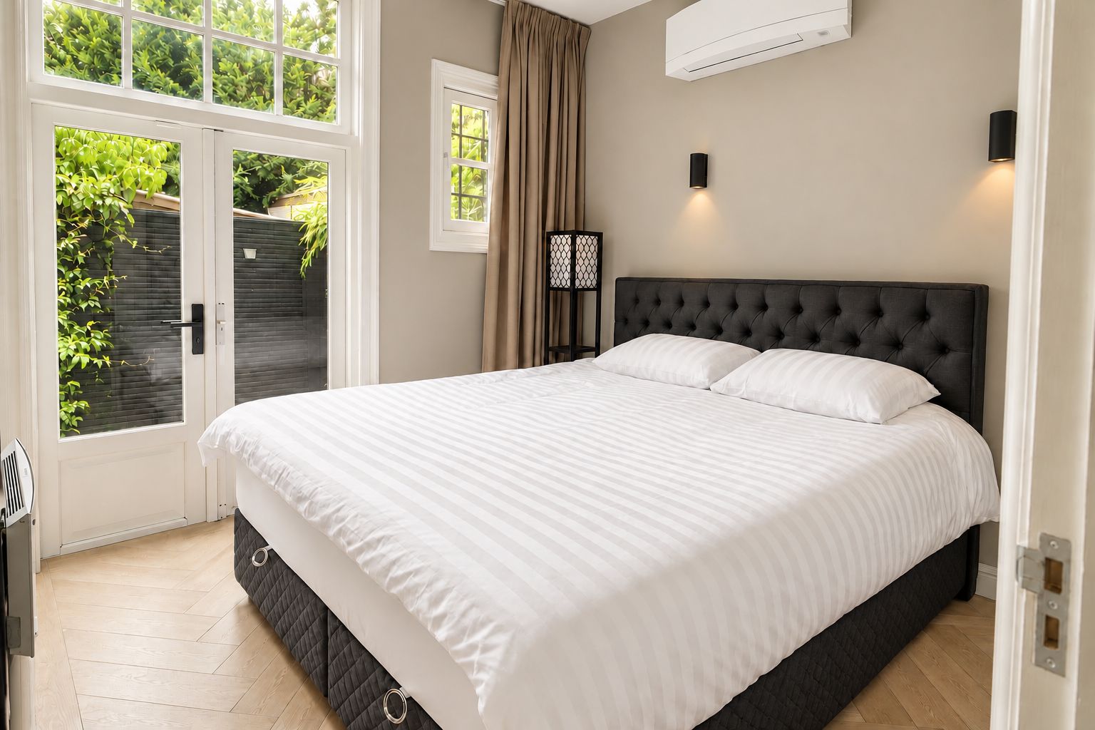 A bedroom with a large bed, a window with curtains, an air conditioning unit, and a wall-mounted light fixture.