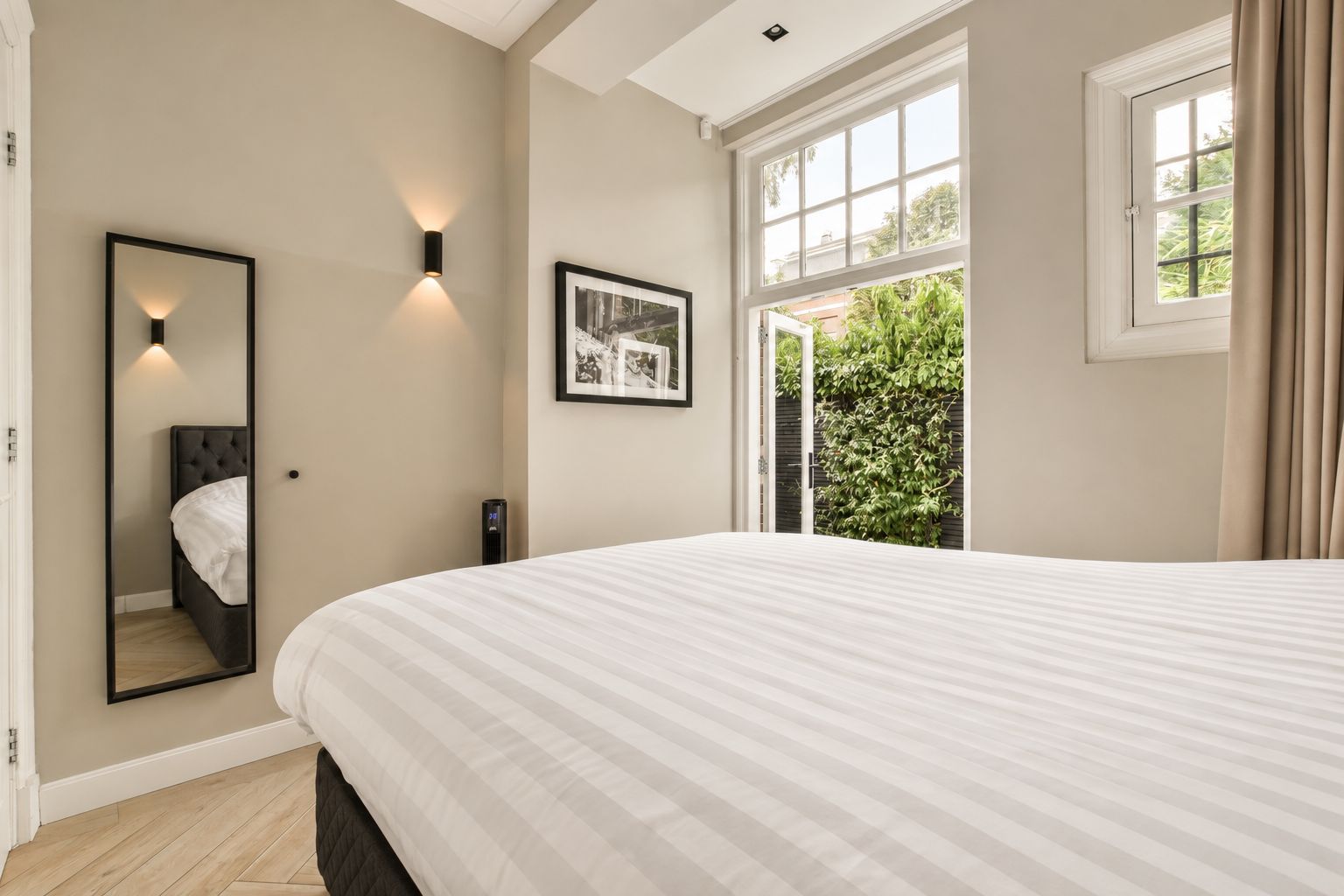 A bedroom with a large bed, a bedside mirror, a wall-mounted light, and a window with curtains.