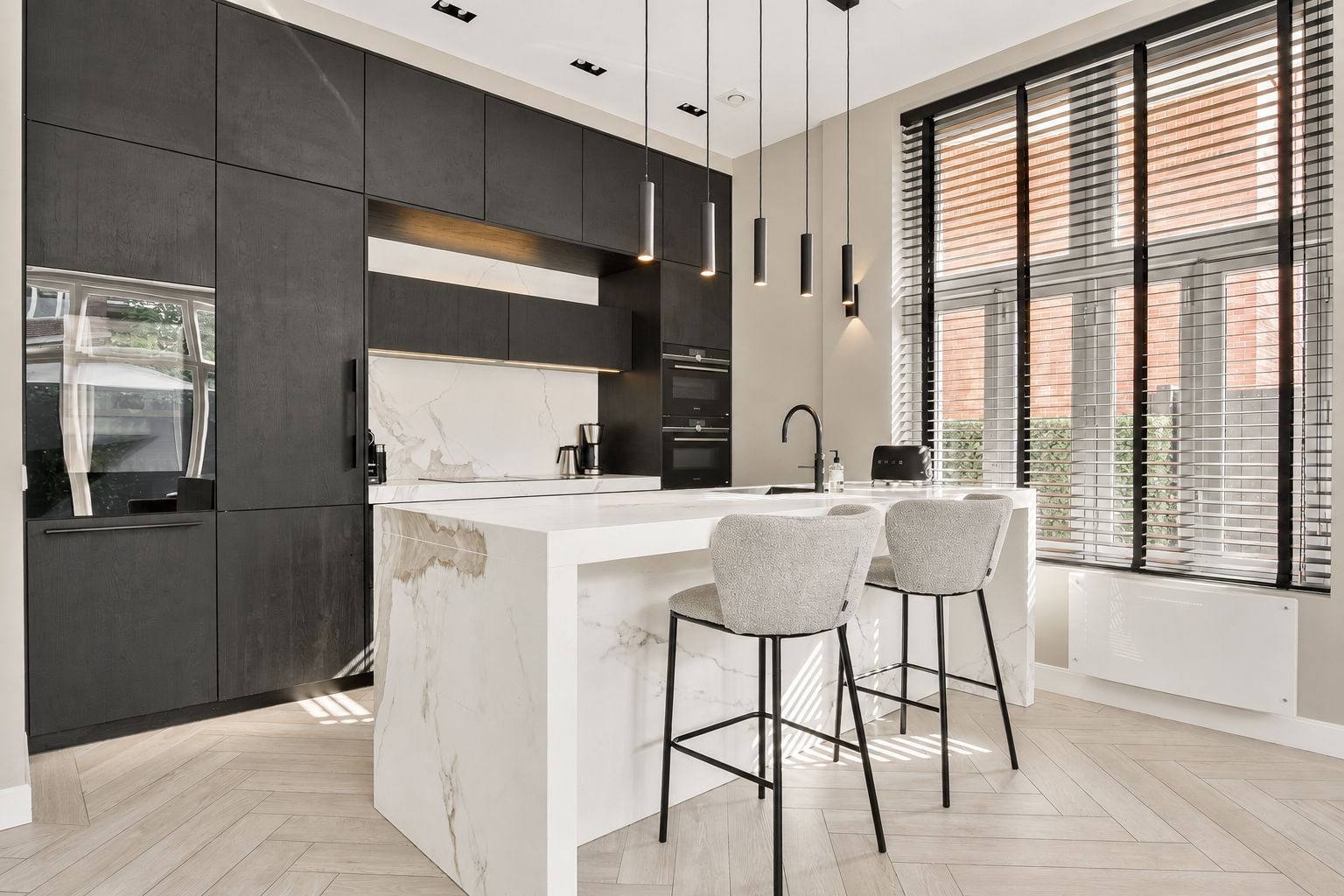 Une cuisine moderne avec des meubles de couleur sombre, un îlot en marbre, des luminaires suspendus et une grande fenêtre équipée de stores.