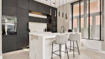 Une cuisine moderne avec des meubles de couleur sombre, un îlot en marbre, des luminaires suspendus et une grande fenêtre équipée de stores.