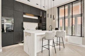 A modern kitchen with dark cabinetry, a marble island, pendant lighting, and a large window with blinds.