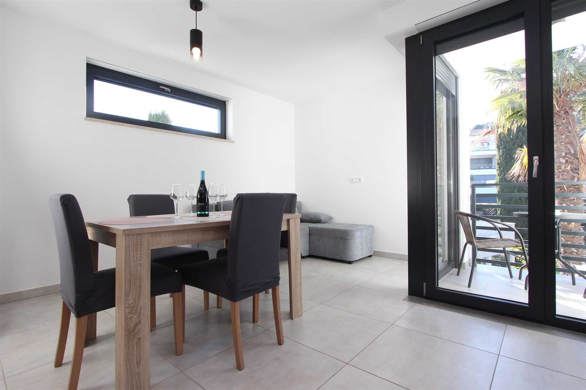 A dining area with a wooden table and four chairs, a bottle of wine, and a window with a view of the outdoors.