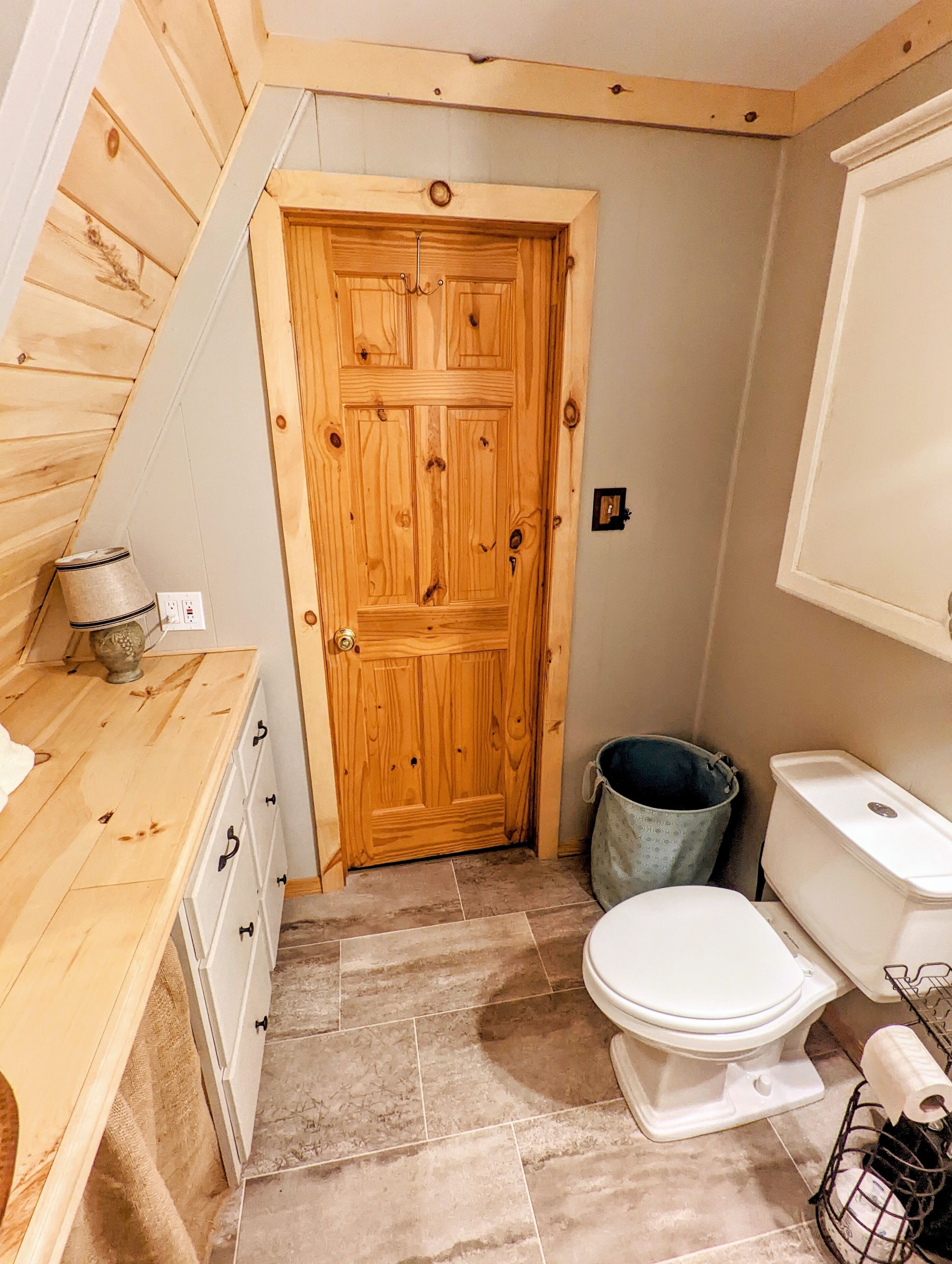 Een houten deur met een hendel, een wit toilet, een afvalbak en een houten toonbank met lades.